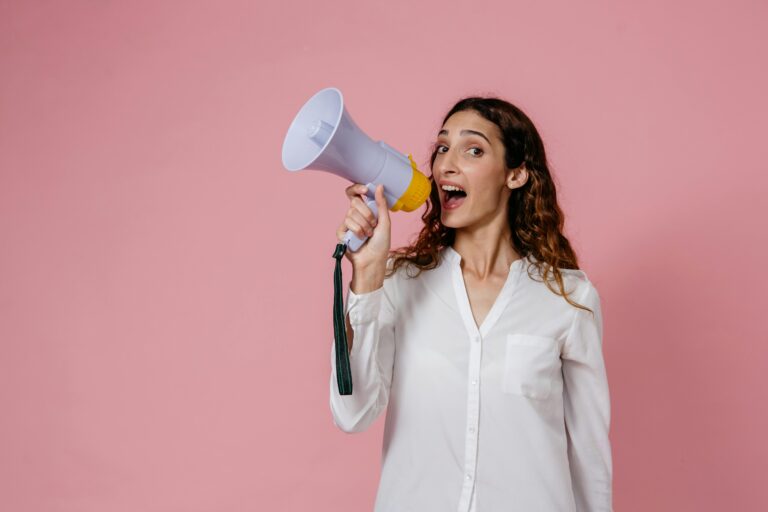 Woman in white shirt holding megaphone against a pink background, confidently speaking.